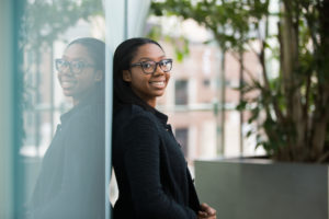 Girl in glasses and blazer smiles and leans up against reflective window with tree in background