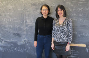 Two women stand in front of chalk board covered in equations.