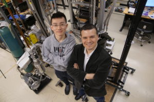 two men stand in a lab with crossed arms, looking up at the camera overhead