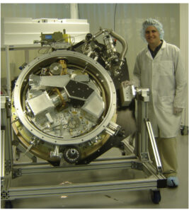 Professor Robert Simcoe, who is wearing a white lab coat, a plastic hair cap, and latex gloves, stands next to the infrared spectrometer FIRE.