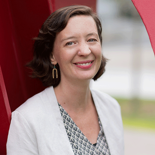 Headshot of a woman standing outside in white blazer and checkered shirt.