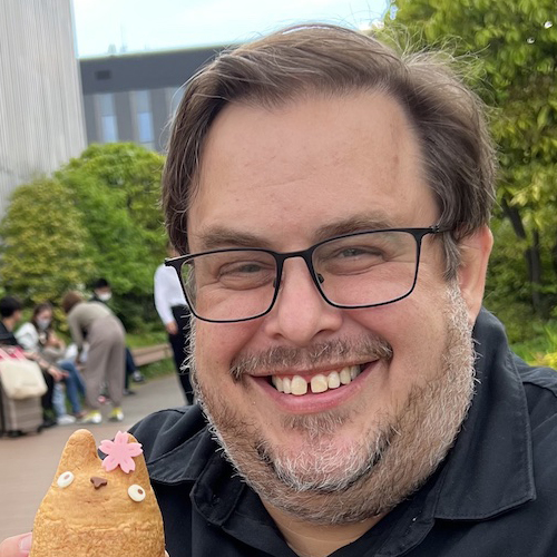 close up of white man with beard and glasses smiling at the camera holding a cat-shaped cookie