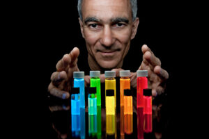 Moungi Bawendi's face and hands to float above and behind two rows of brightly-colored vials containing blue, green, yellow, orange, and red quantum dots.