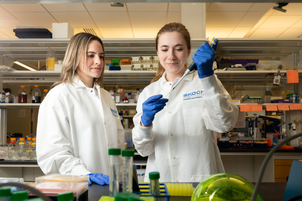 Yadira Soto-Feliciano and Renee Barbosa in a lab wearing white Koch Institute coats. Renee is holding a pipette.