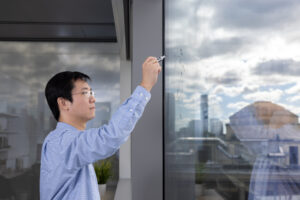 Kaiming He writes on a window with a marker. Clouds and the city skyline are visible.