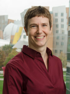 A shite woman with short dark hair and a red buttondown shirt stands inside her lab overlooking Hockfield court.