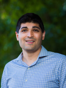 A man with dark hair and a blue buttondown shirt stands outside