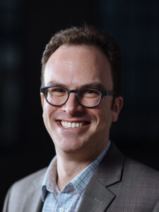 Headshot of a white man with glasses in a gray blazer and blue buttondown shirt